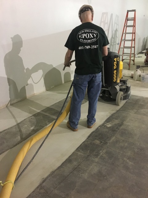 A worker uses a PDG 8000 floor grinder and vacuum hose on a concrete surface, preparing it for epoxy flooring installation.
