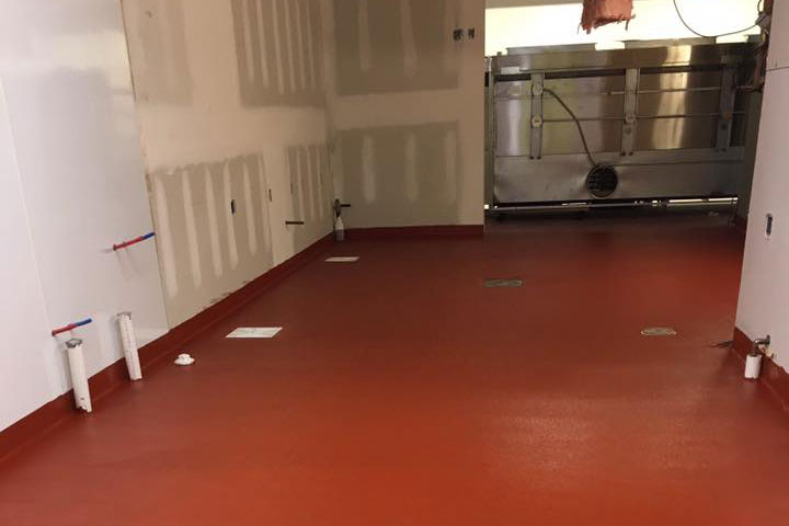 Red epoxy-coated floor in a commercial kitchen under construction. Visible plumbing fixtures and stainless steel equipment in the background.