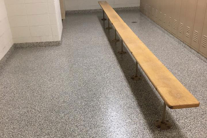 Wooden bench in a locker room with speckled gray flooring. Lockers line one wall, providing storage for athletes or gym users.