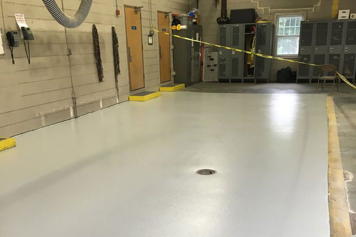 Freshly coated gray epoxy flooring in a garage, showcasing a clean surface and yellow safety markings. Storage lockers and equipment are visible in the background.