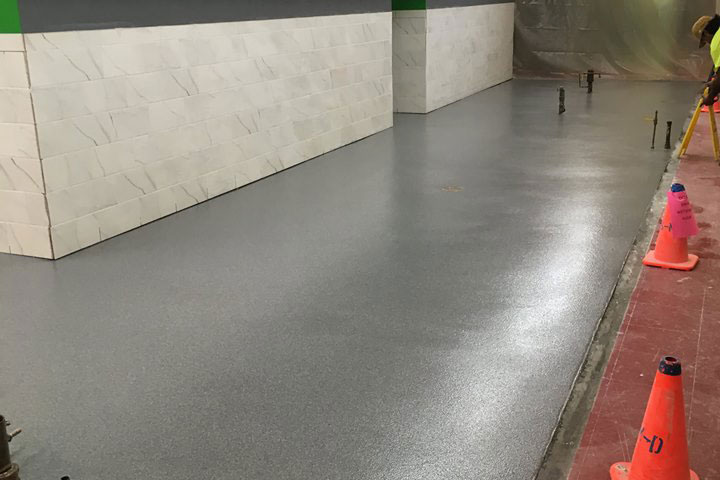 Freshly poured gray flooring in a construction area, with white tiled walls in the background. Safety cones signal ongoing work.